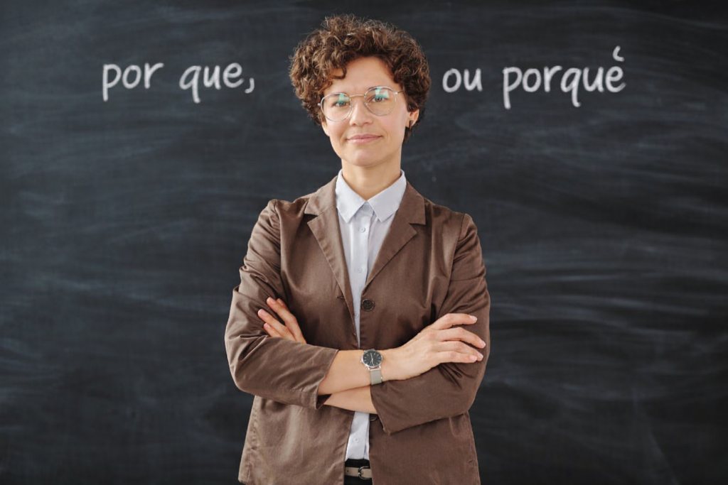Professora de pé na frente de um quadro-negro com as palavras 'por que' e 'porque' escrito, explicando a diferença de uso entre esses termos em português.